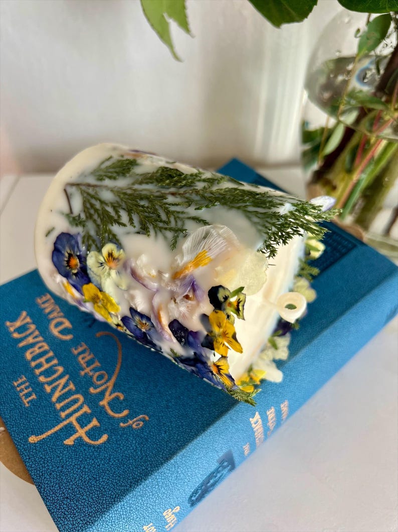 May include: A white candle decorated with pressed flowers and greenery, resting on a blue book titled "The Hunchback of Notre Dame". The candle features yellow, blue, and white floral accents and green sprigs.