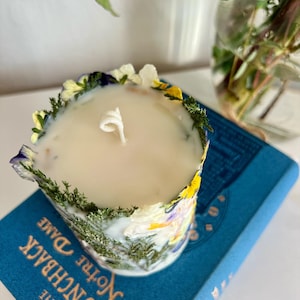 May include: A scented candle in a floral-decorated container. The candle is encircled by dried flowers and greenery. The candle rests on a blue book titled "The Hunchback of Notre Dame".