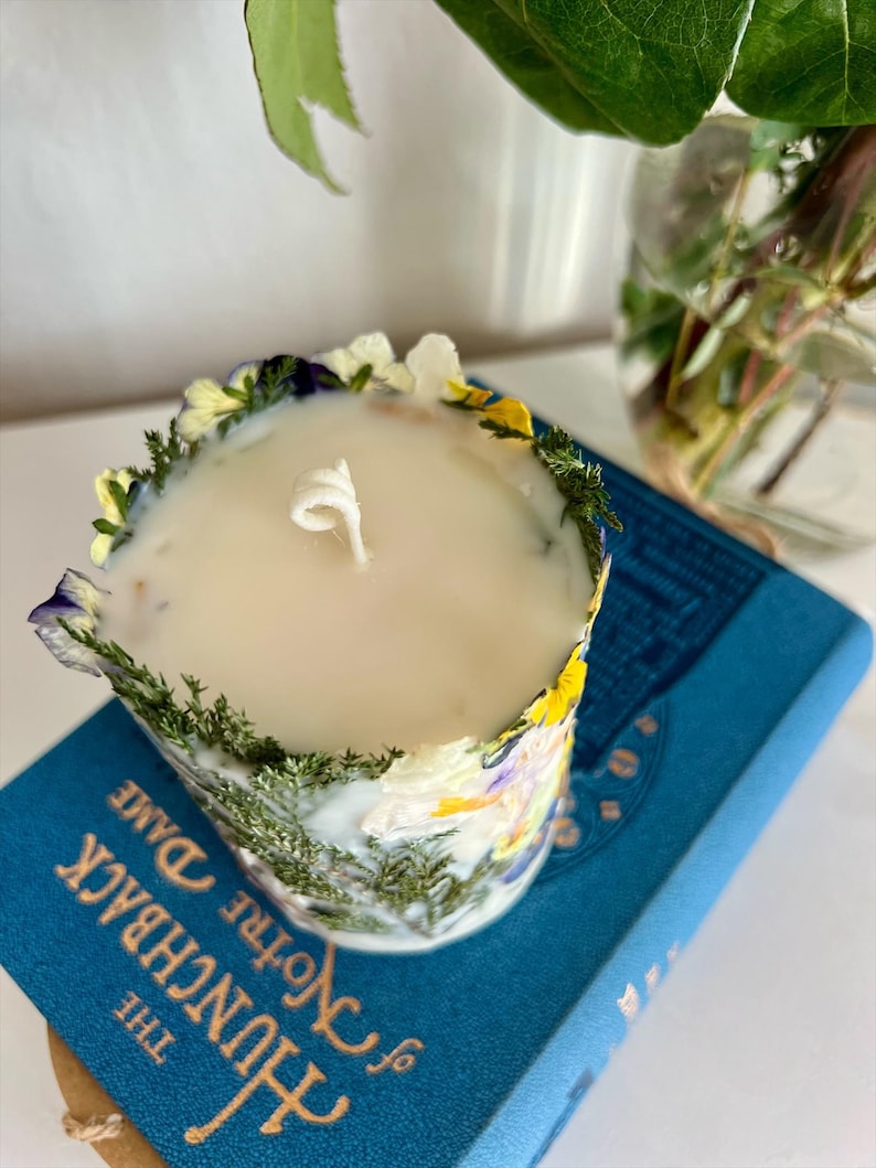 May include: A scented candle in a floral-decorated container. The candle is encircled by dried flowers and greenery. The candle rests on a blue book titled "The Hunchback of Notre Dame".