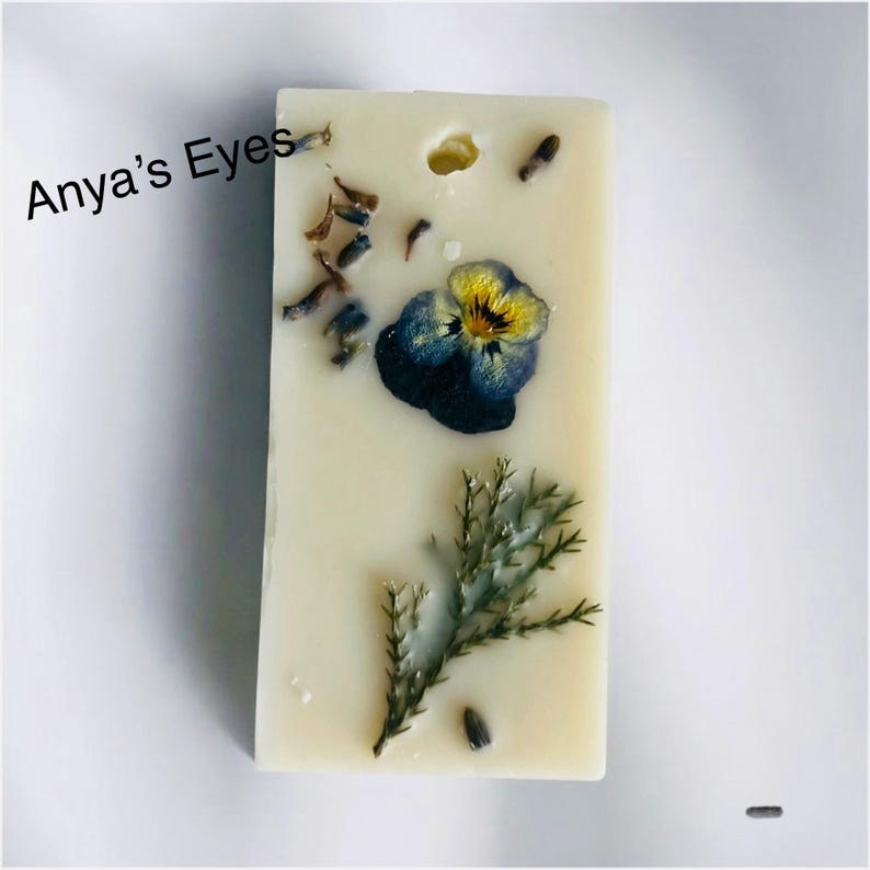 May include: A rectangular, off-white wax melt bar with embedded botanical elements. A blue and yellow pansy, sprigs of greenery, and lavender buds are visible. The text "Anya's Eyes" is printed in black on the upper left corner.