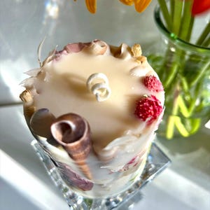 K&ouml;nnte beinhalten: Eine cremefarbene Kerze, verziert mit Muscheln, getrockneten Blumen und kleinen roten Beeren. Die Kerze steht auf einem klaren, quadratischen, facettierten Glassockel. Gelbe und orangefarbene Blumen sind im Hintergrund.