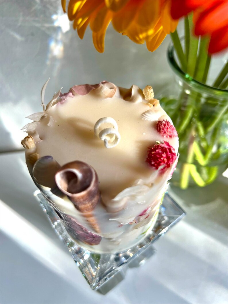 K&ouml;nnte beinhalten: Eine cremefarbene Kerze, verziert mit Muscheln, getrockneten Blumen und kleinen roten Beeren. Die Kerze steht auf einem klaren, quadratischen, facettierten Glassockel. Gelbe und orangefarbene Blumen sind im Hintergrund.