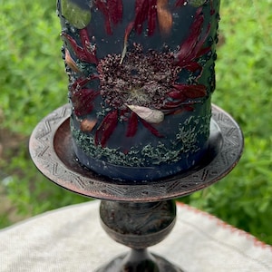 May include: A dark blue pillar candle decorated with dried flowers and greenery, on a decorative bronze candle holder. The candle has a white wick. The candle holder is made of bronze. The background is blurred green.