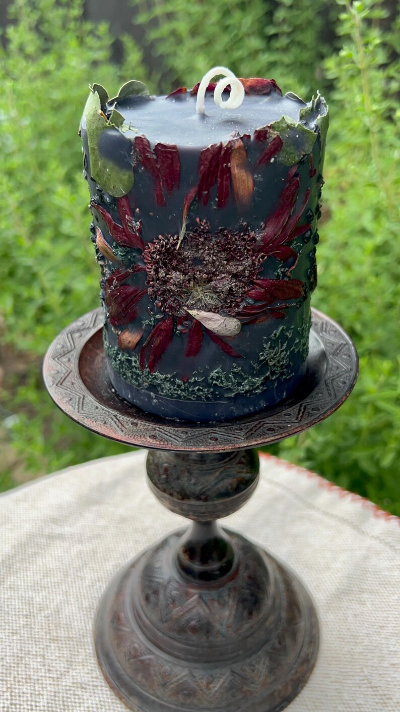 May include: A dark blue pillar candle decorated with dried flowers and greenery, on a decorative bronze candle holder. The candle has a white wick. The candle holder is made of bronze. The background is blurred green.