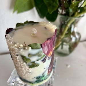 K&ouml;nnte beinhalten: Eine dekorative Kerze mit wei&szlig;em Wachs, verziert mit getrockneten Bl&auml;ttern und Bl&uuml;tenbl&auml;ttern. Die Kerze steht auf einem transparenten, quadratischen St&auml;nder. Eine rosa Rose und gr&uuml;ne Bl&auml;tter sind im Hintergrund zu sehen.