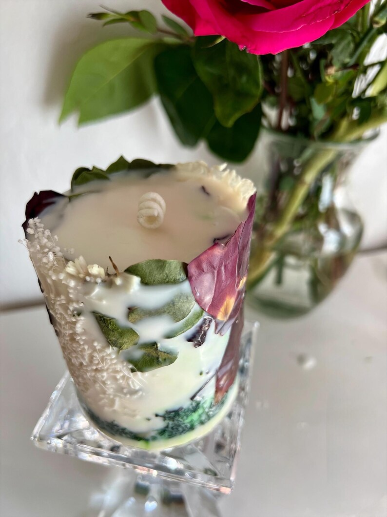 K&ouml;nnte beinhalten: Eine dekorative Kerze mit wei&szlig;em Wachs, verziert mit getrockneten Bl&auml;ttern und Bl&uuml;tenbl&auml;ttern. Die Kerze steht auf einem transparenten, quadratischen St&auml;nder. Eine rosa Rose und gr&uuml;ne Bl&auml;tter sind im Hintergrund zu sehen.