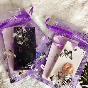 May include: Two small, purple organza bags with silver butterfly accents. One bag contains a dark rectangle with a silver butterfly, the other contains a white rectangle with a dried flower and a silver butterfly. The bags are on a white surface.