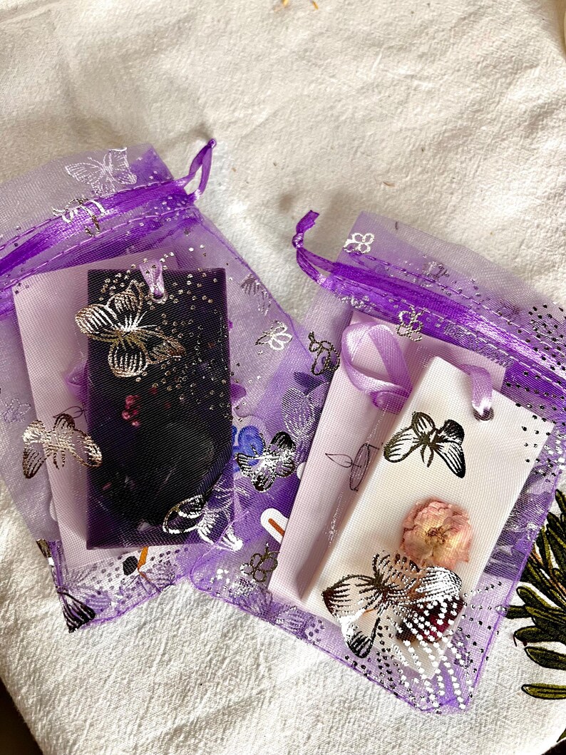 May include: Two small, purple organza bags with silver butterfly accents. One bag contains a dark rectangle with a silver butterfly, the other contains a white rectangle with a dried flower and a silver butterfly. The bags are on a white surface.