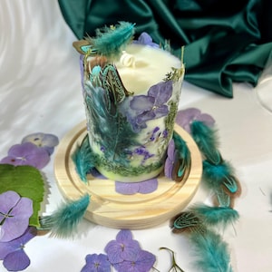 Peut inclure: Une bougie cylindrique en cire blanche orn&eacute;e de p&eacute;tales d'hortensia violets, de mousse verte et de plumes sarcelle incrust&eacute;s. La bougie poss&egrave;de une m&egrave;che blanche et repose sur un socle circulaire en bois clair, entour&eacute;e de p&eacute;tales violets et de plumes.
