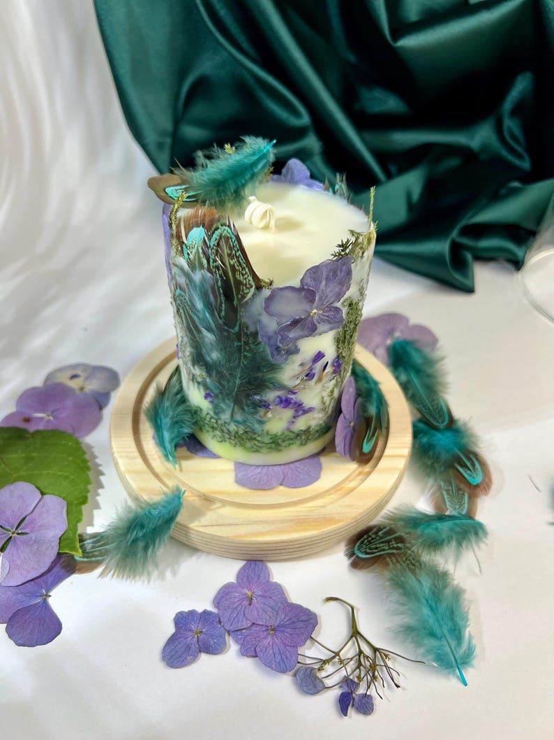 Peut inclure: Une bougie cylindrique en cire blanche orn&eacute;e de p&eacute;tales d'hortensia violets, de mousse verte et de plumes sarcelle incrust&eacute;s. La bougie poss&egrave;de une m&egrave;che blanche et repose sur un socle circulaire en bois clair, entour&eacute;e de p&eacute;tales violets et de plumes.