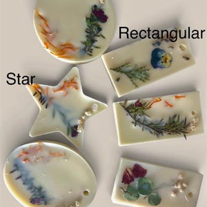 May include: Assortment of cream-colored wax melts in round, rectangular, star, and oval shapes. Each melt is decorated with dried flowers and botanicals. The shapes have small holes for hanging. The word "Round", "Rectangular", "Star", and "Oval" are written on the image.