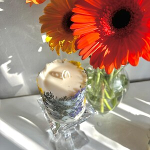 May include: A decorative candle with a floral design, resting on a clear glass stand. The candle is cylindrical, with a cream-colored wax and a wick. The candle's exterior features a botanical pattern with purple and green accents. Bright orange gerbera daisies are in the background.