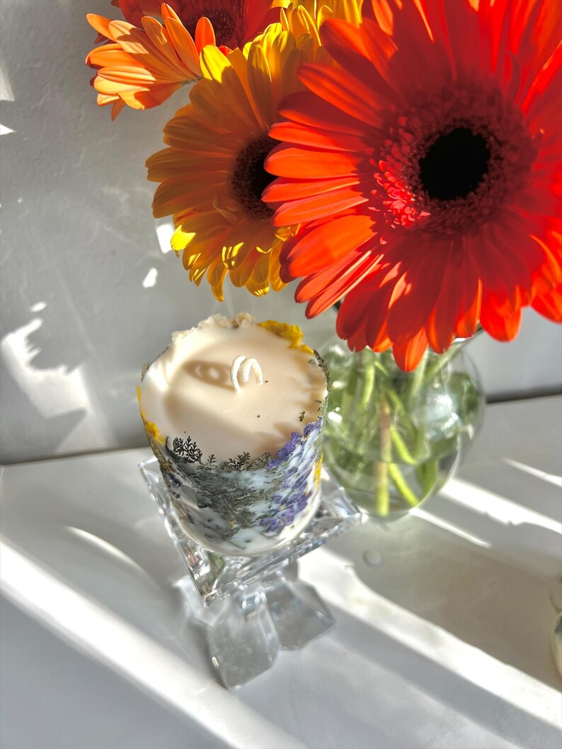 May include: A decorative candle with a floral design, resting on a clear glass stand. The candle is cylindrical, with a cream-colored wax and a wick. The candle's exterior features a botanical pattern with purple and green accents. Bright orange gerbera daisies are in the background.