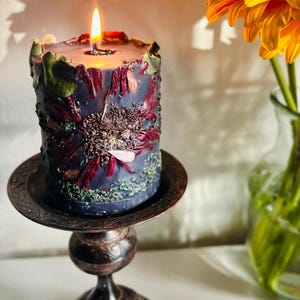 May include: A lit, cylindrical candle in a dark blue colour, decorated with dried flowers in red and green. The candle is on a decorative, antique-style metal stand. A vibrant orange flower in a vase is in the background.
