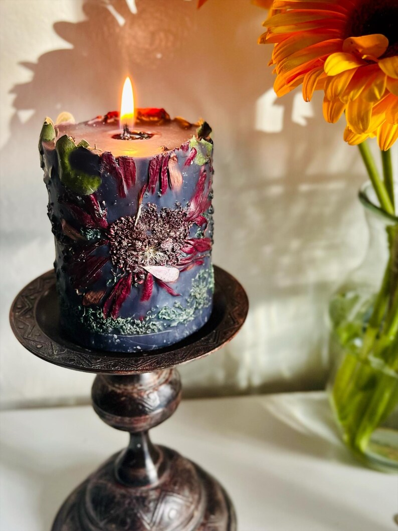 May include: A lit, cylindrical candle in a dark blue colour, decorated with dried flowers in red and green. The candle is on a decorative, antique-style metal stand. A vibrant orange flower in a vase is in the background.