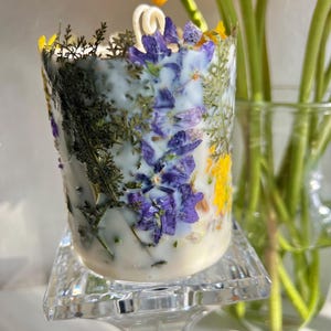 May include: A white candle with embedded dried flowers, including purple, yellow, and green accents. The candle sits on a clear, square-shaped glass stand. A yellow flower and green stems are in the background.