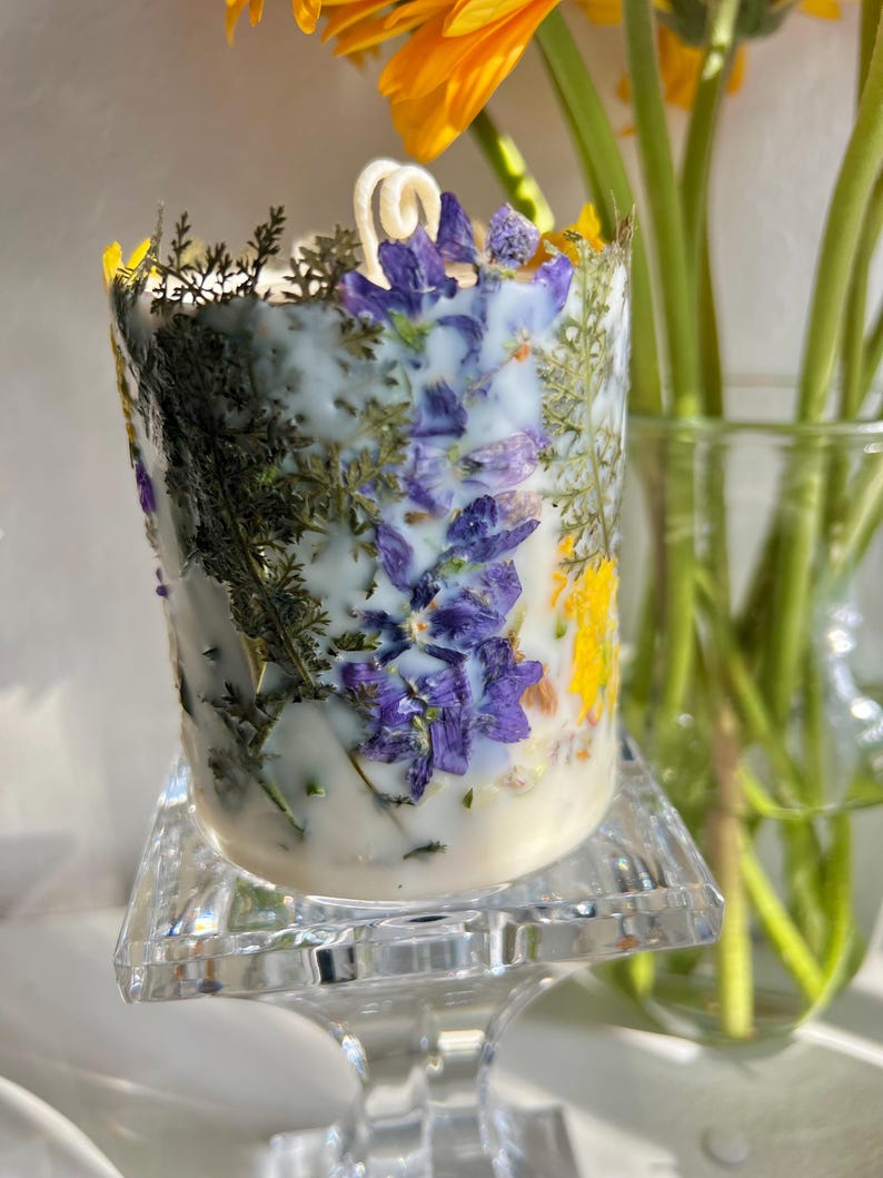 May include: A white candle with embedded dried flowers, including purple, yellow, and green accents. The candle sits on a clear, square-shaped glass stand. A yellow flower and green stems are in the background.