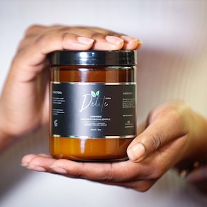 May include: A brown glass jar with a black label that says "Delité Ayurvedic Deep Moisturizing Bountiful" with green leaf icons. The jar is being held by a person's hands.
