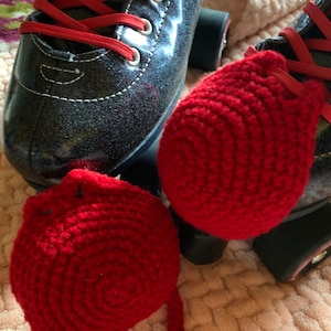 May include: Two red crocheted roller skate stoppers. The stoppers are round and have a small loop on the side. The stoppers are sitting on a pink fuzzy surface.