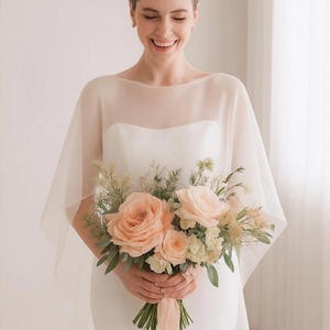 May include: A bride in a white wedding dress and sheer cape smiles, holding a bouquet of peach roses and greenery. The bouquet is tied with a peach ribbon. The background is a soft, neutral color.