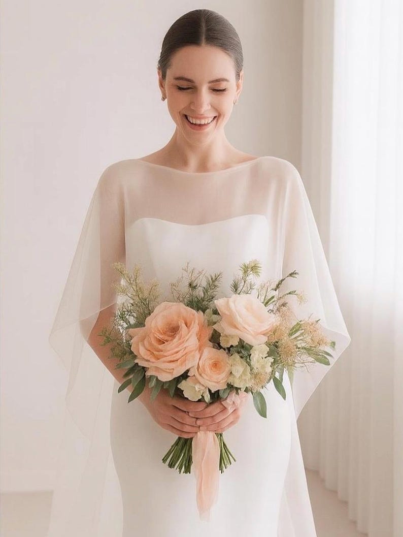 May include: A bride in a white wedding dress and sheer cape smiles, holding a bouquet of peach roses and greenery. The bouquet is tied with a peach ribbon. The background is a soft, neutral color.