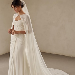 May include: A white wedding dress with a cape and a long train. The dress has a sweetheart neckline and a fitted bodice with draping. The cape flows from the shoulders, adding an elegant touch. The veil is attached to the hair.