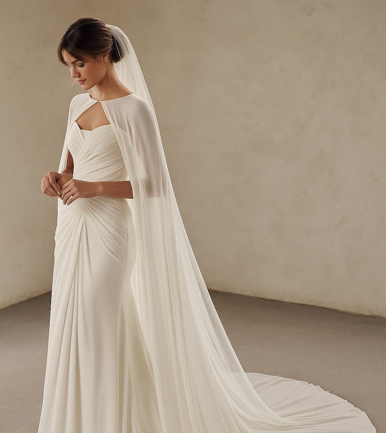 May include: A white wedding dress with a cape and a long train. The dress has a sweetheart neckline and a fitted bodice with draping. The cape flows from the shoulders, adding an elegant touch. The veil is attached to the hair.