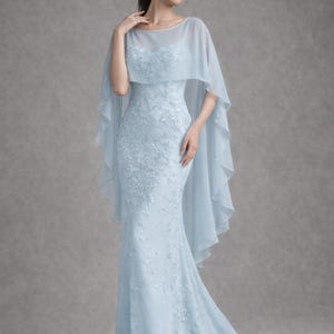 K&ouml;nnte beinhalten: Ein hellblaues, bodenlanges Kleid mit Meerjungfrau-Silhouette. Das Kleid hat aufwendige Blumenstickereien und einen transparenten Umhang. Das Model tr&auml;gt das Kleid in einem Studio.