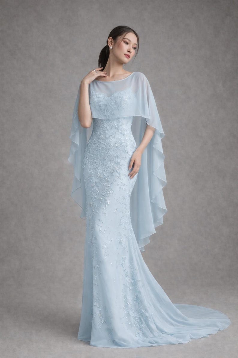 K&ouml;nnte beinhalten: Ein hellblaues, bodenlanges Kleid mit Meerjungfrau-Silhouette. Das Kleid hat aufwendige Blumenstickereien und einen transparenten Umhang. Das Model tr&auml;gt das Kleid in einem Studio.