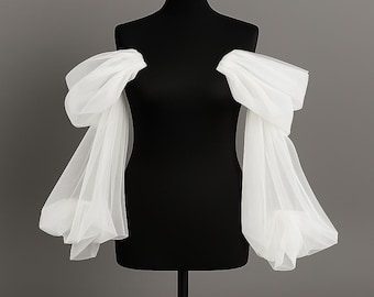 May include: A black dress form displays a pair of white, sheer, off-the-shoulder sleeves. The sleeves are voluminous, with a gathered, puffy design at the shoulder and a flowing, elongated shape down the arm. The background is a solid gray.