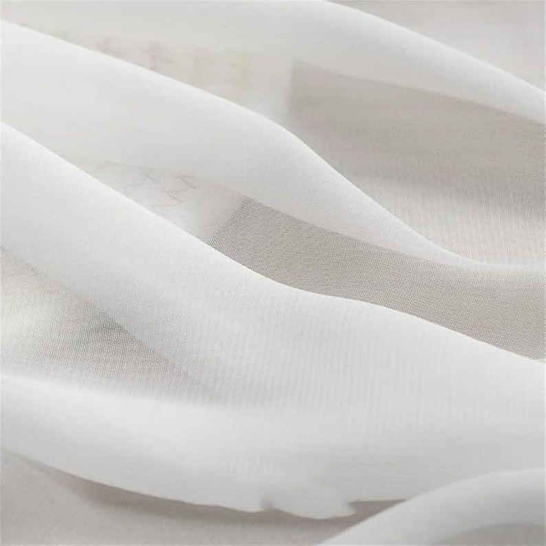 May include: Close-up of white fabric with soft folds and a translucent appearance. The fabric has a subtle texture and a flowing, airy quality. The image highlights the delicate nature of the material, suggesting it could be used for clothing or home decor.