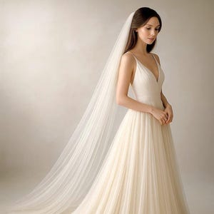 May include: A cream-colored wedding dress with a plunging neckline and a flowing skirt. The dress is paired with a long, sheer veil. The bride is standing in front of a neutral background.