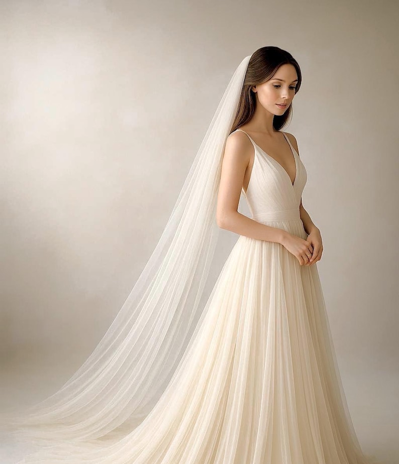 May include: A cream-colored wedding dress with a plunging neckline and a flowing skirt. The dress is paired with a long, sheer veil. The bride is standing in front of a neutral background.