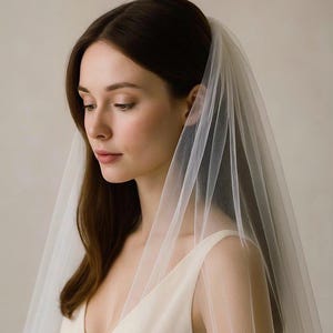 May include: A woman wearing a sheer, white bridal veil. The veil is draped over the head and shoulders, cascading down the front. The woman is wearing a cream-colored dress with a deep V-neckline. The background is a soft, neutral tone.