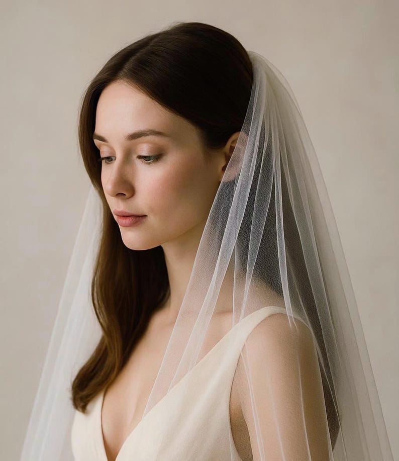 May include: A woman wearing a sheer, white bridal veil. The veil is draped over the head and shoulders, cascading down the front. The woman is wearing a cream-colored dress with a deep V-neckline. The background is a soft, neutral tone.