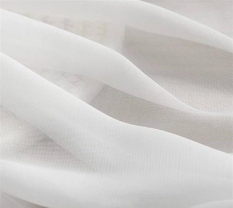 May include: Close-up of white fabric with soft folds and a translucent appearance. The fabric has a delicate texture and a subtle sheen, creating a sense of movement and flow. The image highlights the fabric's lightness and airy quality.