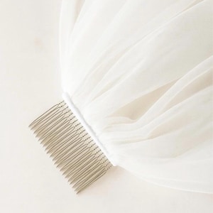 May include: A white bridal veil with a comb attached. The veil is made of a sheer, flowing fabric, and the comb is silver-toned. The fabric is gathered at the comb, creating soft folds.