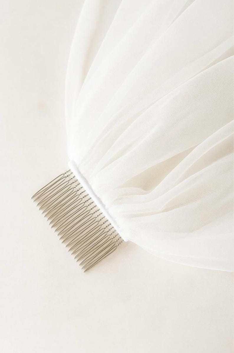 May include: A white bridal veil with a comb attached. The veil is made of a sheer, flowing fabric, and the comb is silver-toned. The fabric is gathered at the comb, creating soft folds.