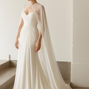 May include: A white wedding dress with a sweetheart neckline and a flowing skirt. The dress features a sheer cape that drapes over the shoulders. The dress is made of a soft, flowing fabric and has a train.