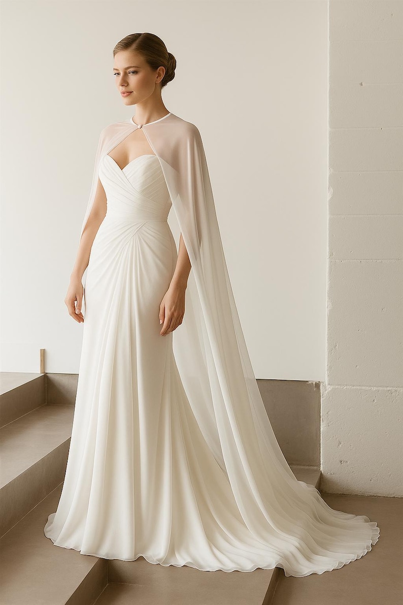 May include: A white wedding dress with a sweetheart neckline and a flowing skirt. The dress features a sheer cape that drapes over the shoulders. The dress is made of a soft, flowing fabric and has a train.