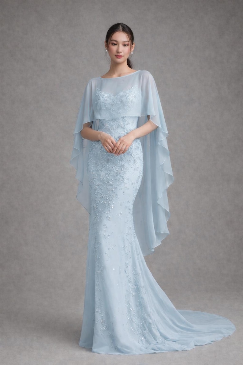 K&ouml;nnte beinhalten: Ein hellblaues, bodenlanges Kleid mit Meerjungfrau-Silhouette. Das Kleid hat zarte Blumenstickereien und einen transparenten Umhang. Das Kleid eignet sich f&uuml;r formelle Anl&auml;sse wie Hochzeiten oder Galas.
