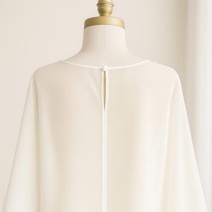 May include: Ivory-colored, flowing garment on a mannequin. The back of the top features a keyhole opening with a white trim. The neckline is also trimmed in white. The fabric drapes softly, suggesting a lightweight material.