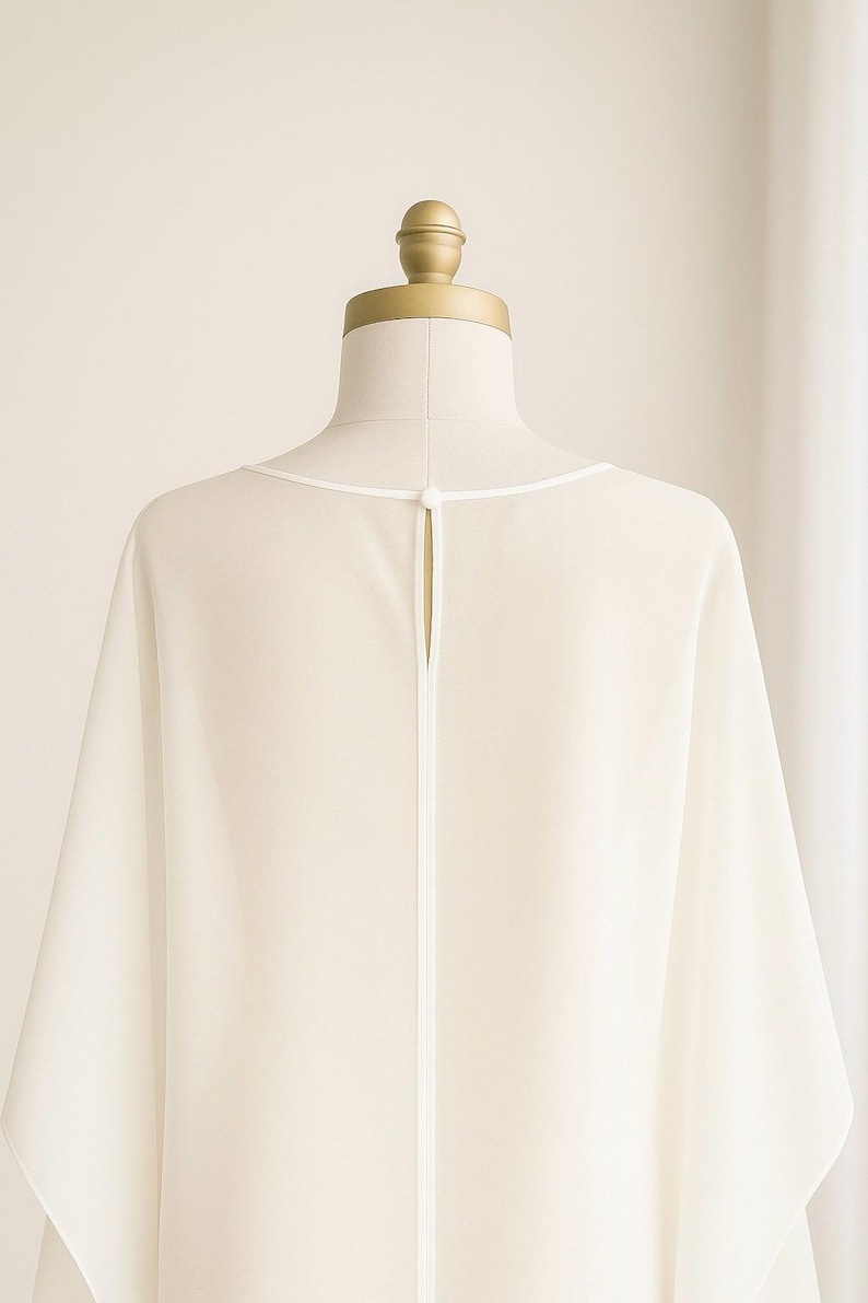 May include: Ivory-colored, flowing garment on a mannequin. The back of the top features a keyhole opening with a white trim. The neckline is also trimmed in white. The fabric drapes softly, suggesting a lightweight material.