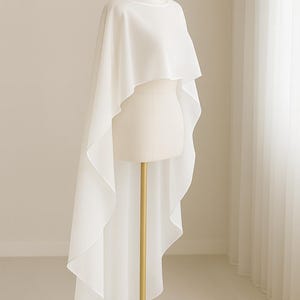 May include: A white, flowing cape-style garment on a mannequin. The fabric drapes elegantly, with a longer back hem. The mannequin is on a gold stand with a round base, set against a neutral background.