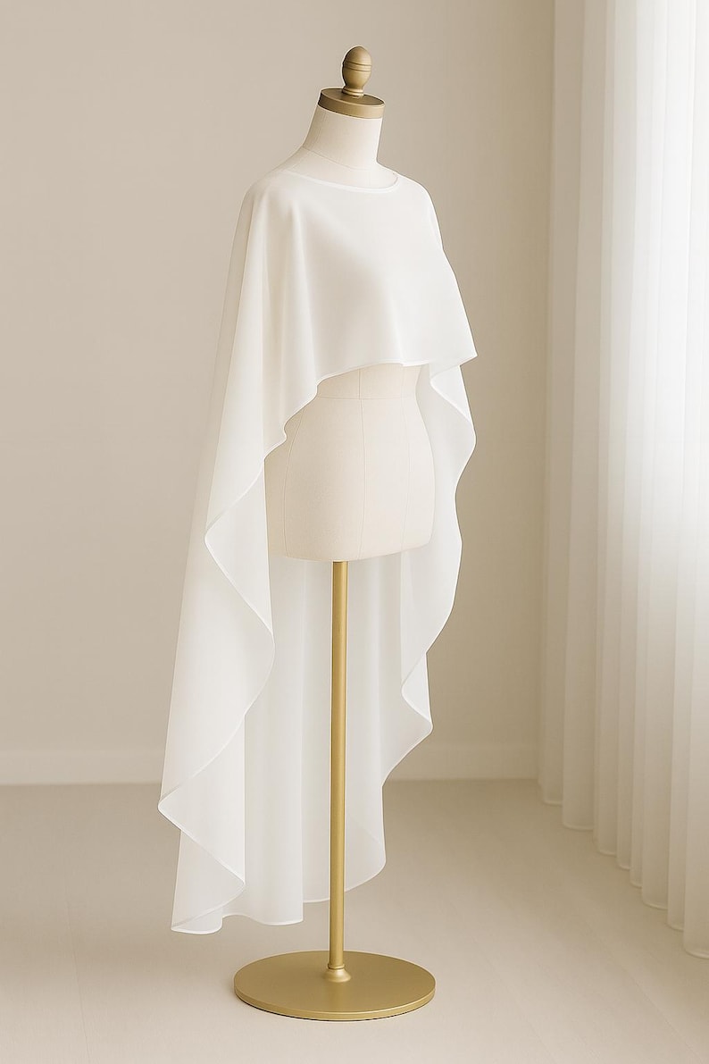 May include: A white, flowing cape-style garment on a mannequin. The fabric drapes elegantly, with a longer back hem. The mannequin is on a gold stand with a round base, set against a neutral background.