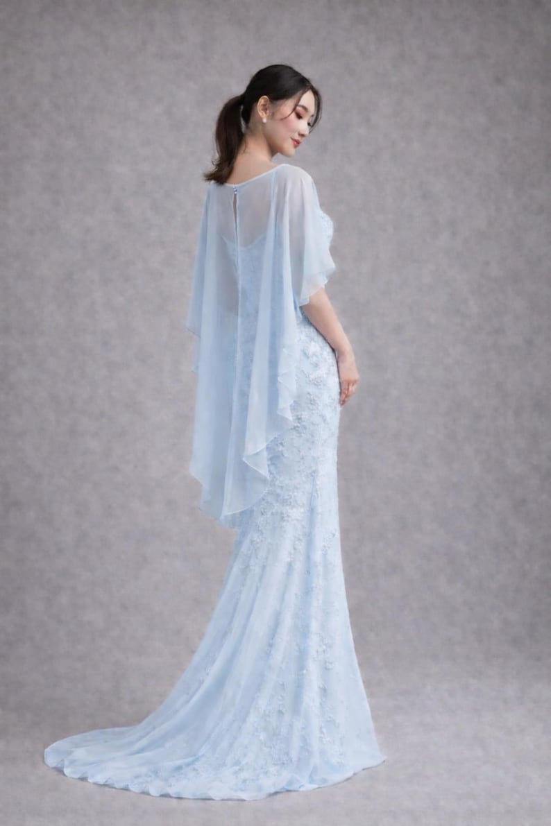 K&ouml;nnte beinhalten: Ein hellblaues, bodenlanges Kleid mit aufwendigen Spitzendetails. Das Kleid hat eine figurbetonte Silhouette, die sich am Saum ausstellt. Ein durchsichtiges, flie&szlig;endes Capelet bedeckt die Schultern und verleiht einen eleganten Touch. Das Kleid ist f&uuml;r formelle Anl&auml;sse geeignet.