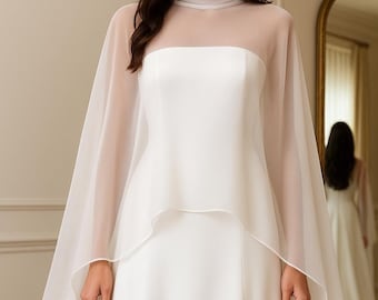 May include: A white bridal gown with a sheer cape overlay. The dress features a high neckline and a fitted bodice. The cape drapes over the shoulders and arms, creating a flowing silhouette. The gown is elegant and suitable for a wedding.