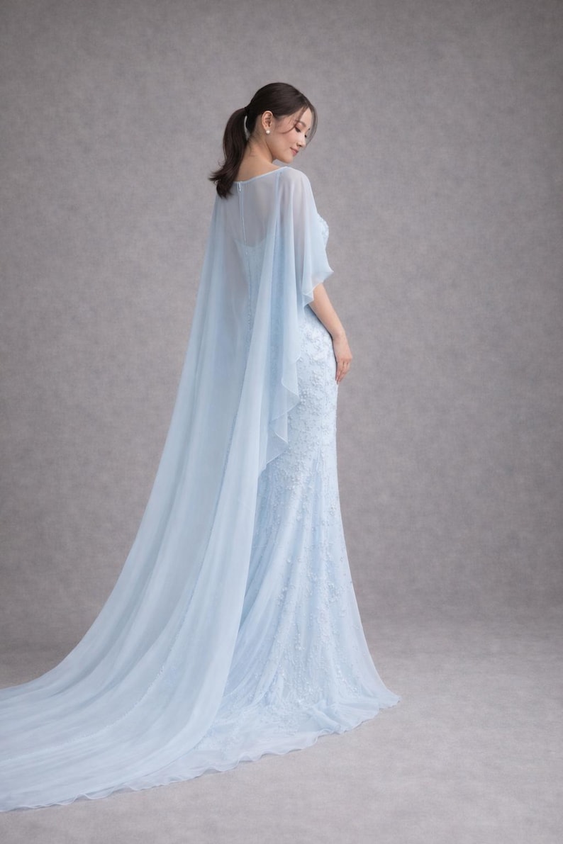 K&ouml;nnte beinhalten: Ein hellblaues, bodenlanges Kleid mit einem flie&szlig;enden Umhang. Das Kleid hat aufwendige Spitzendetails und eine figurbetonte Silhouette. Der Umhang f&auml;llt elegant von den Schultern und erzeugt einen weichen, &auml;therischen Look. Der Stoff wirkt leicht und luftig.