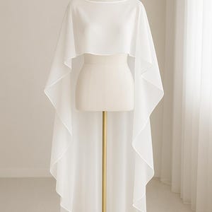 May include: A white, flowing cape-style garment with a boat neckline is displayed on a mannequin. The fabric drapes elegantly, creating a layered effect. The cape is floor-length, with a rounded hemline. The mannequin is on a gold stand.