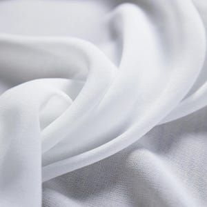 May include: Close-up of flowing white fabric with soft folds and a delicate texture. The fabric appears to be lightweight, possibly silk or chiffon, with a subtle sheen. The image highlights the fabric's smooth surface and gentle curves.